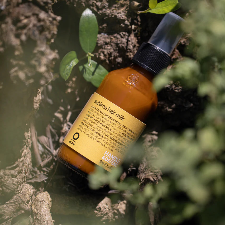 A brown Oway Sublime Hair Milk bottle with a black spray cap lies on textured bark among greenery—a lightweight leave-in conditioner for softness, shine, and frizz control.
