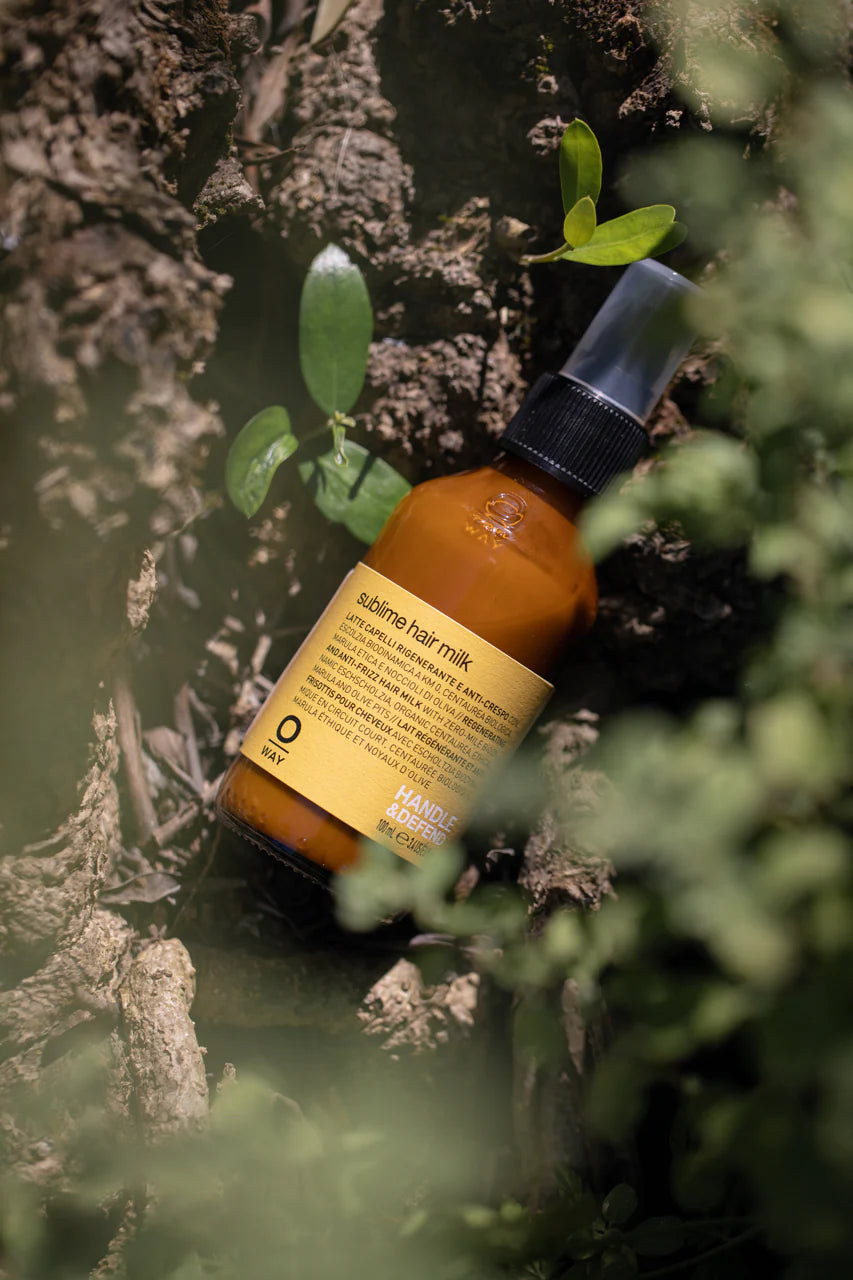 A brown Oway Sublime Hair Milk bottle with a black spray cap lies on textured bark among greenery—a lightweight leave-in conditioner for softness, shine, and frizz control.