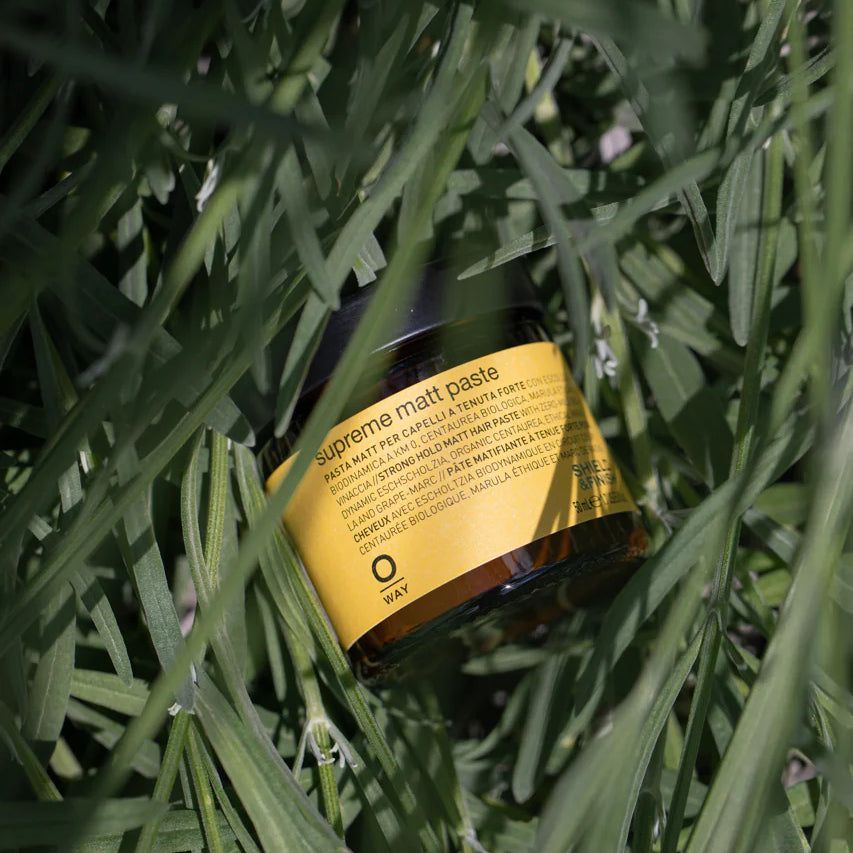 A jar of Oway Supreme Matte Paste, labeled with yellow and partially hidden by tall green grass, suggests its strong hold styling power and healthy matte finish.