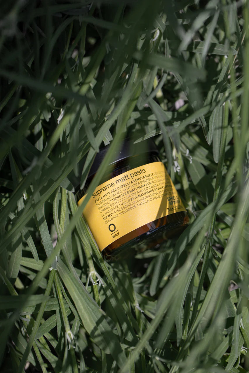 A jar of Oway Supreme Matte Paste, labeled with yellow and partially hidden by tall green grass, suggests its strong hold styling power and healthy matte finish.