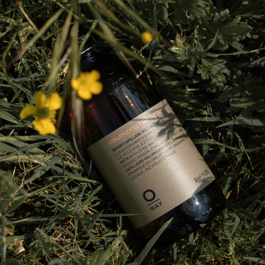 A bottle of Oway Volumizing Hair Bath, a gentle volumizing shampoo for fine or limp hair by Oway, with a beige label rests on green grass beside yellow wildflowers.