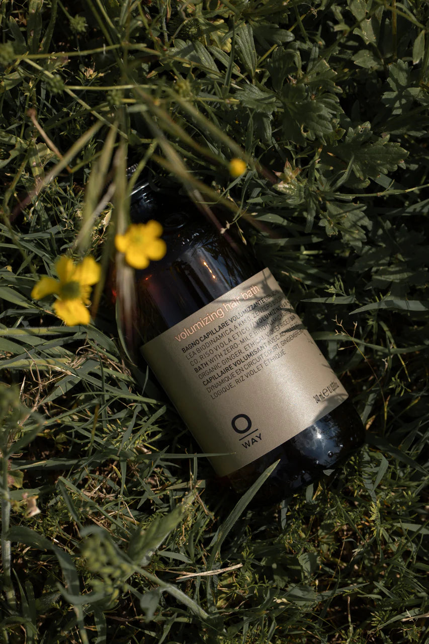 A bottle of Oway Volumizing Hair Bath, a gentle volumizing shampoo for fine or limp hair by Oway, with a beige label rests on green grass beside yellow wildflowers.