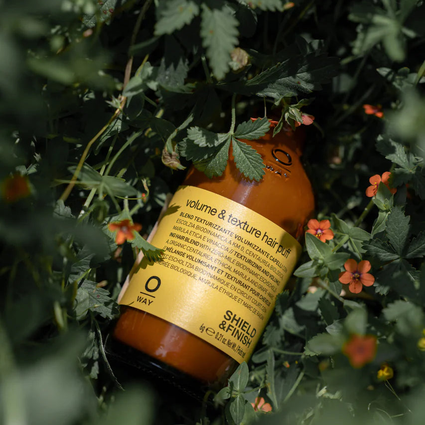 Oway Volume and Texture Hair Puff, a volumizing styling powder for instant lift by Oway, rests on green foliage with small orange flowers, promising fuller hair for fine types.