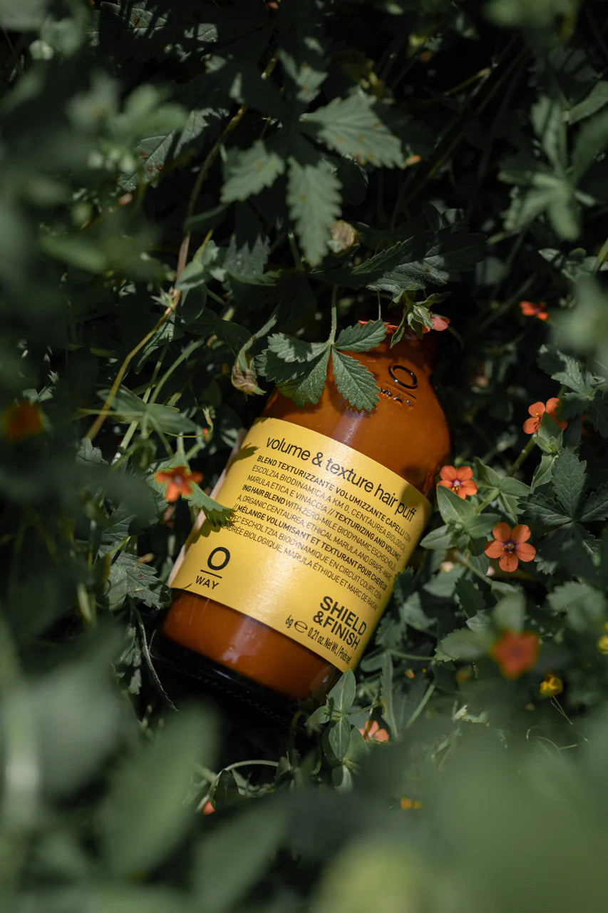 Oway Volume and Texture Hair Puff, a volumizing styling powder for instant lift by Oway, rests on green foliage with small orange flowers, promising fuller hair for fine types.