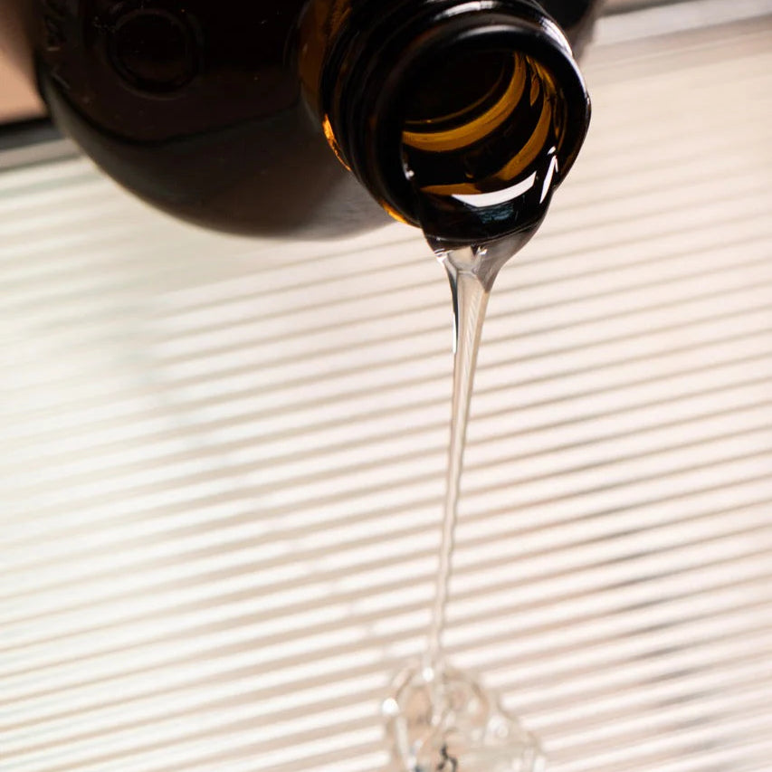 A thick, clear liquid from a dark brown glass bottle of Oway Volumizing Hair Bath—Oway’s gentle volumizing shampoo for fine or limp hair—is being poured onto a striped surface.
