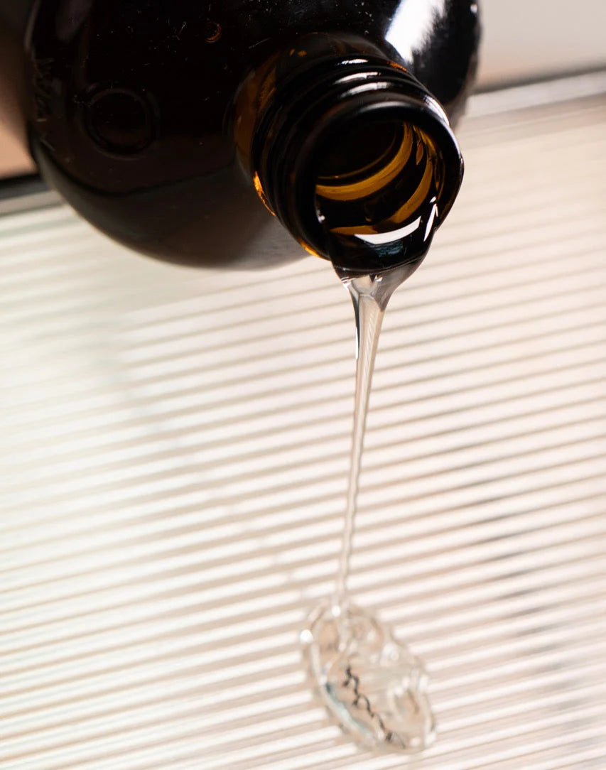 A thick, clear liquid from a dark brown glass bottle of Oway Volumizing Hair Bath—Oway’s gentle volumizing shampoo for fine or limp hair—is being poured onto a striped surface.