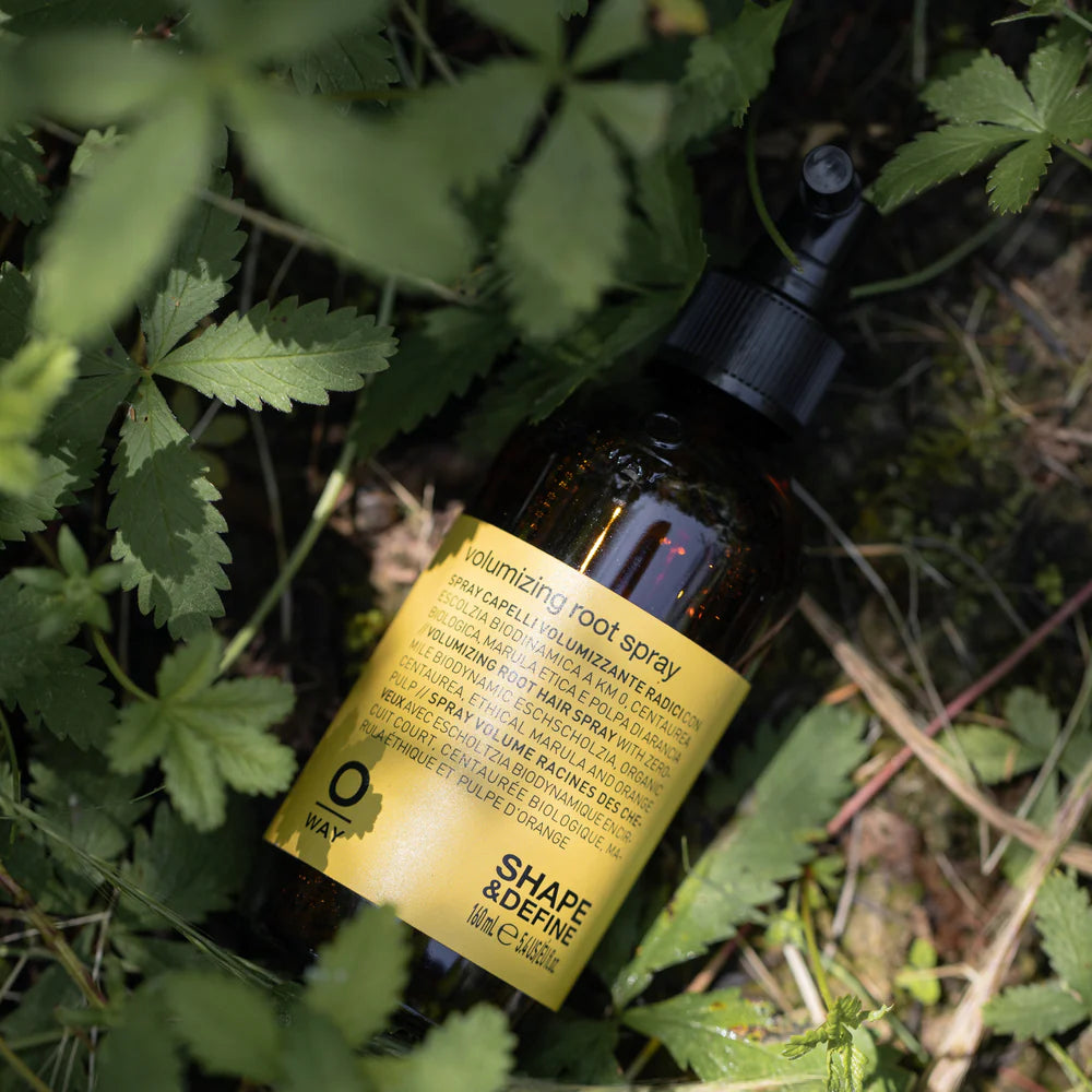 A bottle of Oway Volumizing Root Spray | Lightweight Root Lift for Fine, Flat Hair with a yellow label rests on the ground among green leaves and grass.