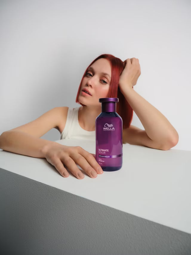 Woman with red hair holding a purple Wella bottle on a gray surface