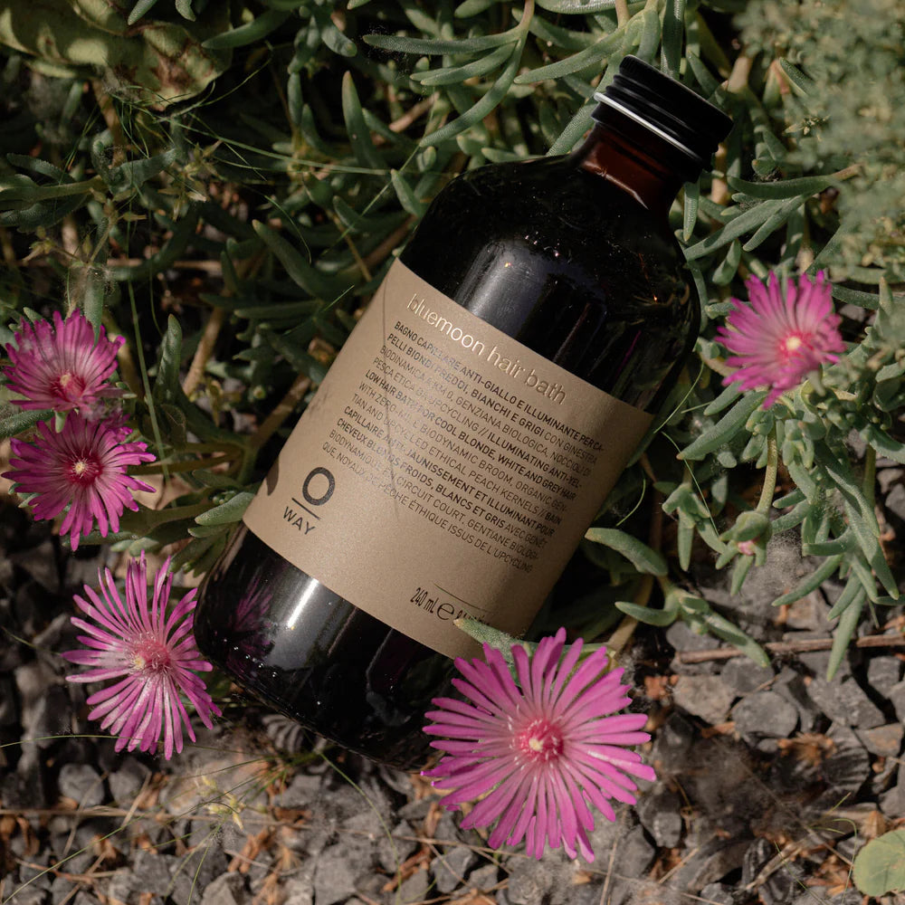 A dark glass bottle labeled "Oway Bluemoon Hair Bath" rests among green plants and pink flowers—an illuminating anti-yellow shampoo by Oway, designed to neutralize brassiness for cool blonde, white, and grey hair.