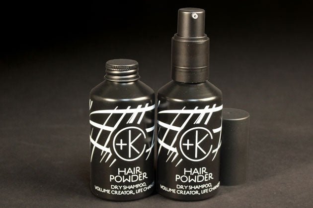 Two black containers of the Cult and King PUMPABLE HAIR POWDER, a talc-free dry shampoo, beautifully presented with antioxidant-rich powder and adorned with white abstract designs alongside a "+K" logo. One container is equipped with a spray nozzle while the other features a screw cap, all elegantly positioned against a black background.