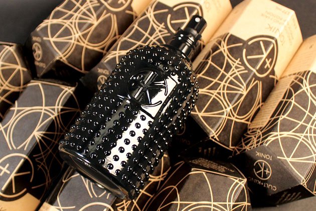 A black, studded perfume bottle lies on a pile of brown and black paper packaging adorned with circular and geometric designs, next to a Cult and King TONIK | Conditioning Hair & Scalp Potion by Cult and King.