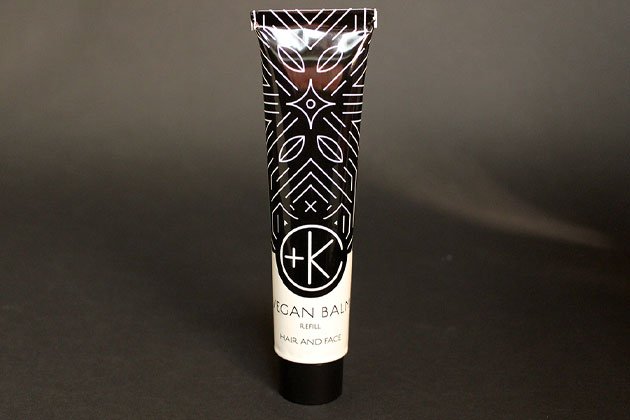 A tube of Cult and King VEGAN BALM against a black background showcases a sleek geometric design. The text reads "Vegan Balm Refill Hair and Face" with a "+K" logo, infused with nourishing botanical oils for hair styling and skin care.