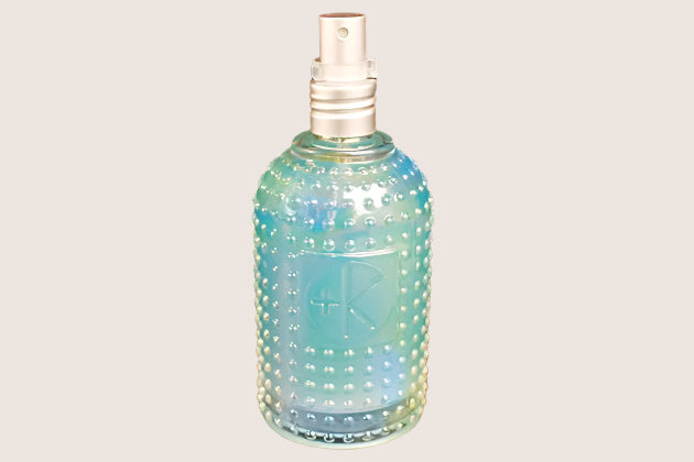 The Cult and King SET SPRAY, a Semi-Precious Tri-Phase Wonderworking Hairspray by Cult and King, comes in a blue-green glass bottle with a silver spray nozzle and a textured, dotted surface. The front of the bottle is embossed with a logo featuring a plus sign. Infused with Malachite Extract, this formulation also provides heat protection. The background is black.