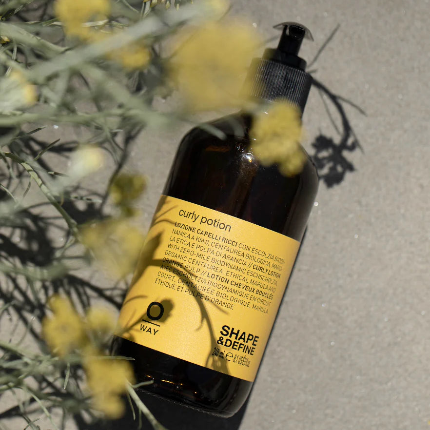 A brown bottle labeled "Oway Curly Potion | Curl-Defining Serum for Soft, Shiny Curls" rests on sand beside yellow flowers and green foliage, highlighting Oway's organic serum that enhances and defines curls.