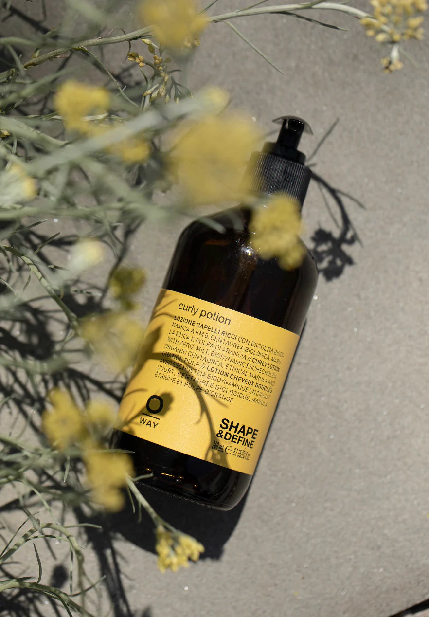 A brown bottle labeled "Oway Curly Potion | Curl-Defining Serum for Soft, Shiny Curls" rests on sand beside yellow flowers and green foliage, highlighting Oway's organic serum that enhances and defines curls.