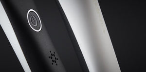Close-up of the GHD PLATINUM+ STYLER 1" FLAT IRON by GHD, a black and white device with a power button and small speaker grille, featuring Ultra-zone™ predictive technology, shown against a dark background.