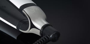 Close-up of the GHD PLATINUM+ STYLER - 1" FLAT IRON, showing its sleek metallic exterior, branded design, power cord connection, and advanced Ultra-zone™ predictive technology.