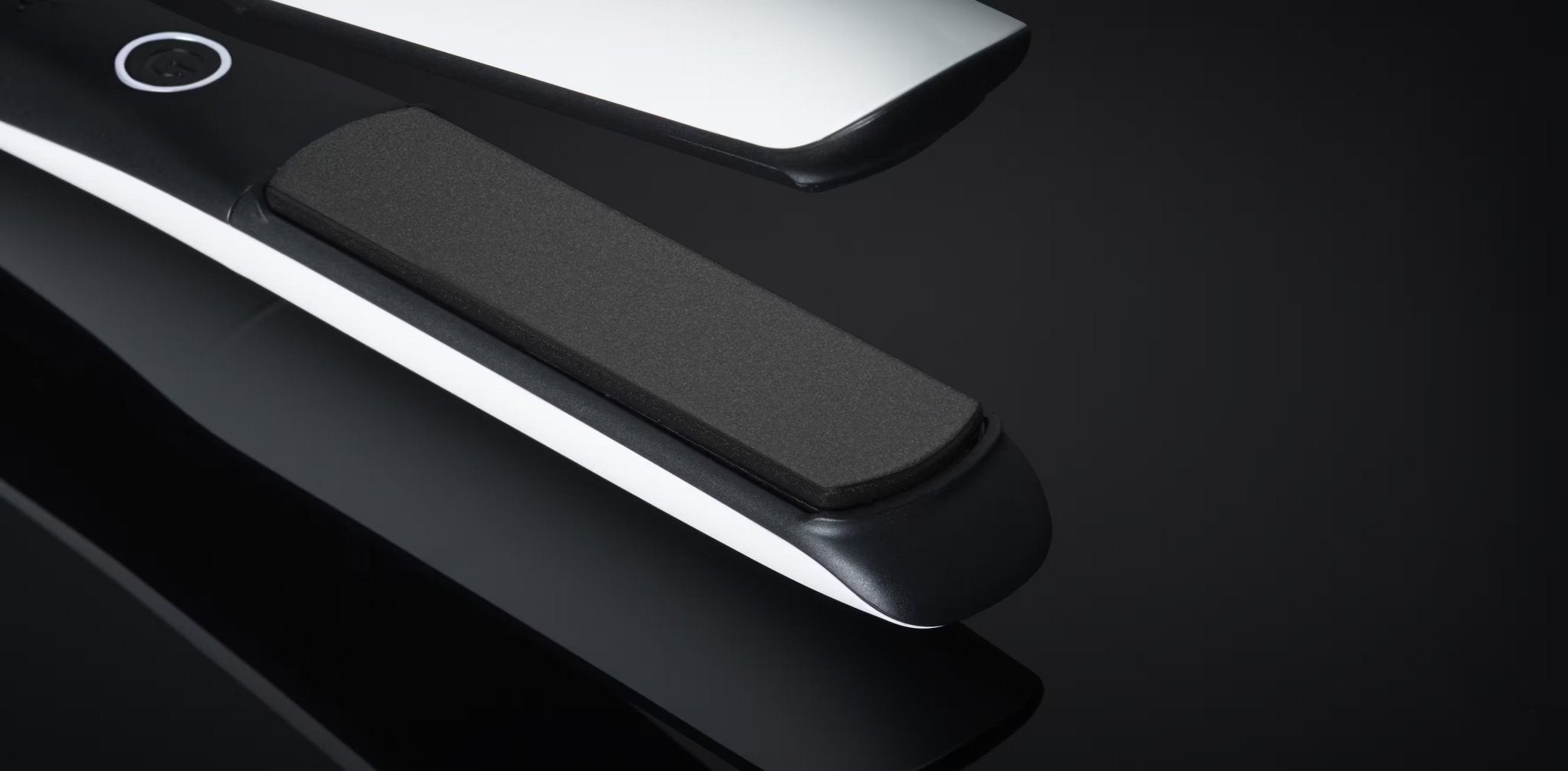 Close-up of a black and white GHD PLATINUM+ STYLER - 1" FLAT IRON with ceramic plates, placed against a reflective black surface.