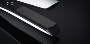 Close-up of the GHD PLATINUM+ STYLER 1" flat iron in black and white, featuring Ultra-zone™ predictive technology, shown on a dark reflective surface.