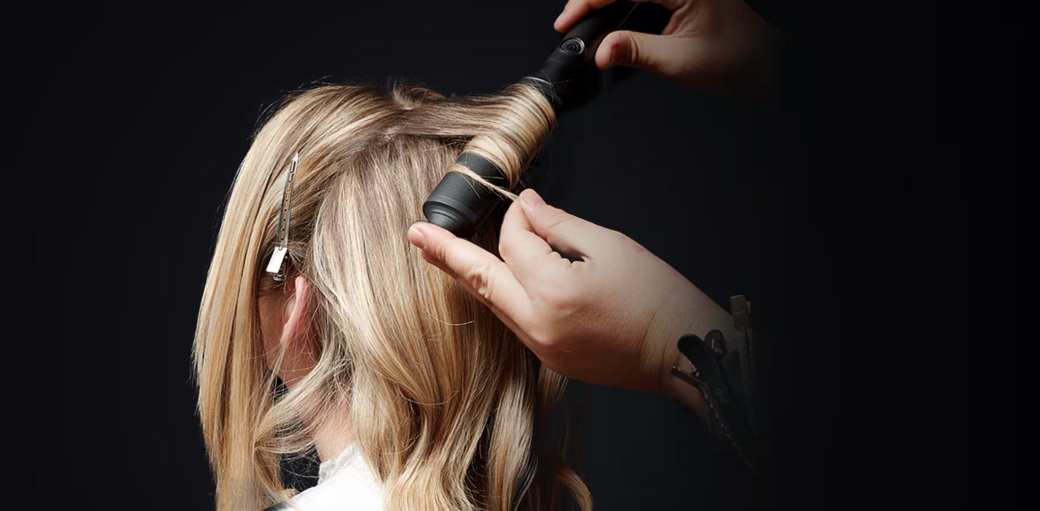 Someone uses the GHD CLASSIC WAVE - OVAL CURLING WAND by GHD to curl blonde hair, with the person sitting and their back to the camera against a dark background.