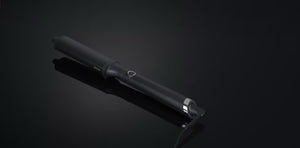 The GHD CLASSIC WAVE - OVAL CURLING WAND by GHD features a ceramic-coated barrel and power button, ideal for creating Hollywood waves, shown here on a dark reflective surface.