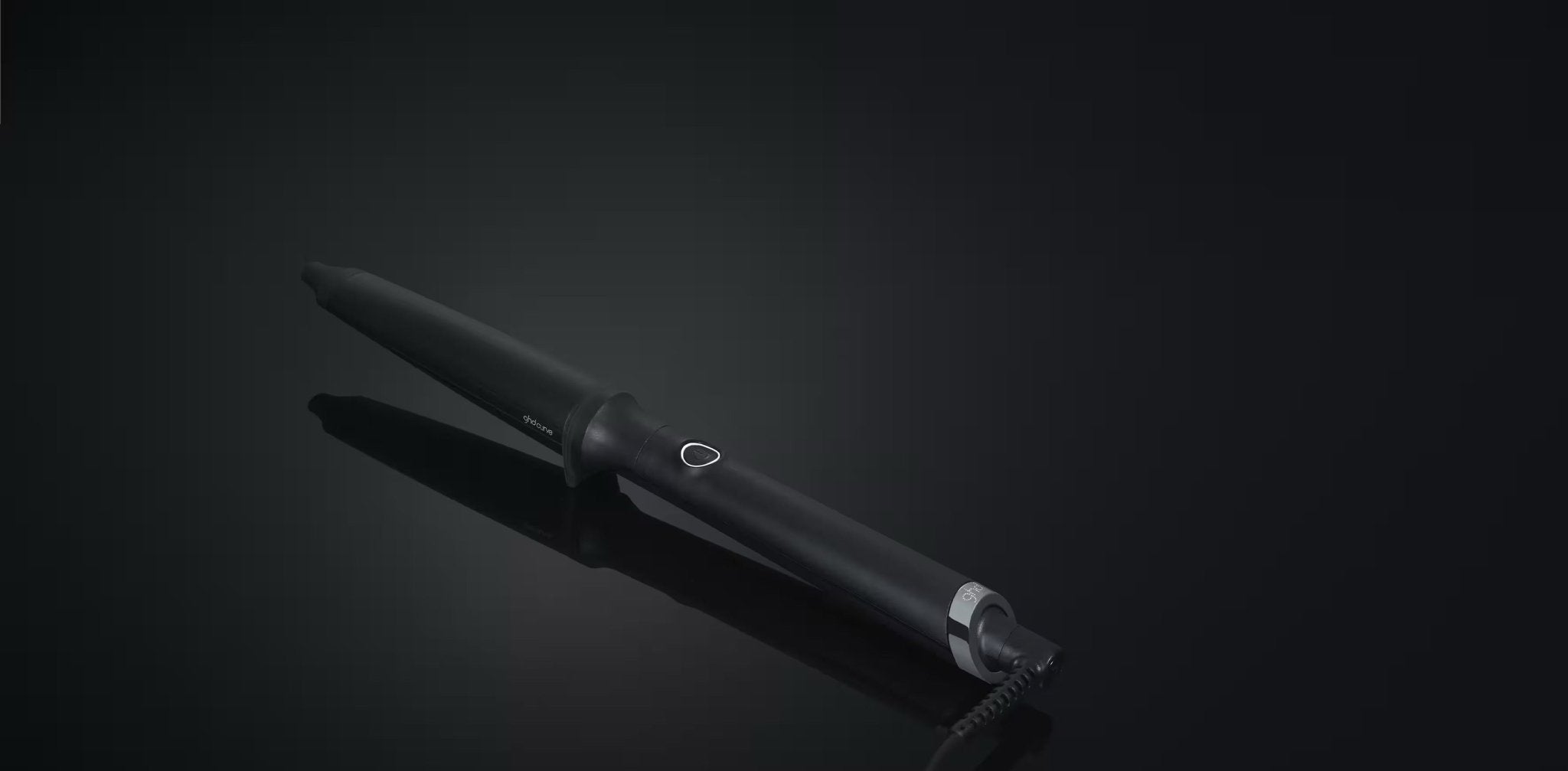 The GHD CREATIVE CURL - TAPERED CURLING WAND IN BLACK by GHD features a matte finish, circular power button, and metal band near the cord—ideal for effortless beachy waves. It's shown resting on a glossy black surface.