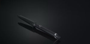The GHD CREATIVE CURL - TAPERED CURLING WAND IN BLACK by GHD features a matte finish, circular power button, and metal band near the cord—ideal for effortless beachy waves. It's shown resting on a glossy black surface.