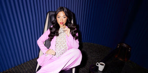 A woman with long, beachy waves styled with the GHD CREATIVE CURL Tapered Curling Wand in Black by GHD sits on a chair against a blue ribbed wall, wearing a light pink suit and holding eyeglasses. A white cup and brown bag are beside her.