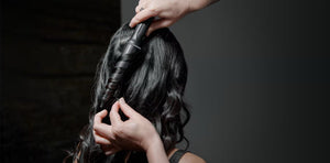 Hands use the GHD CREATIVE CURL - TAPERED CURLING WAND IN BLACK by GHD to curl long, dark hair. The image highlights the back of the head and curling process, creating soft beachy waves against a neutral dark background.