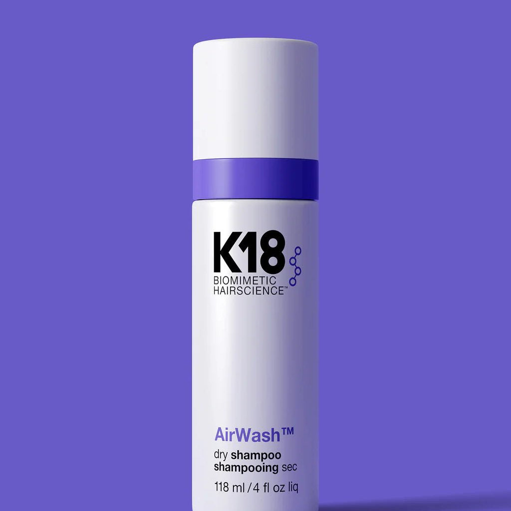 A cylindrical white bottle with a purple stripe, labeled "K18 AirWash Dry Shampoo," stands against a solid purple background. Enhanced with odorBIND™ biotechnology, the bottle holds 118 ml (4 fl oz) and promotes scalp health.