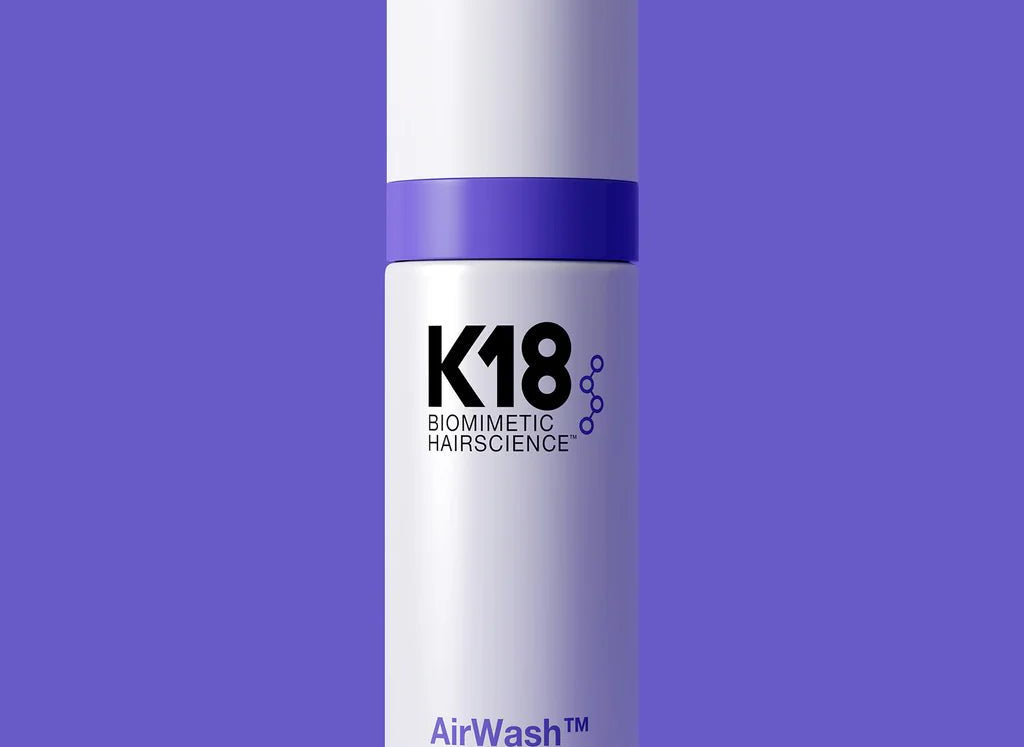 A cylindrical white bottle with a purple stripe, labeled "K18 AirWash Dry Shampoo," stands against a solid purple background. Enhanced with odorBIND™ biotechnology, the bottle holds 118 ml (4 fl oz) and promotes scalp health.