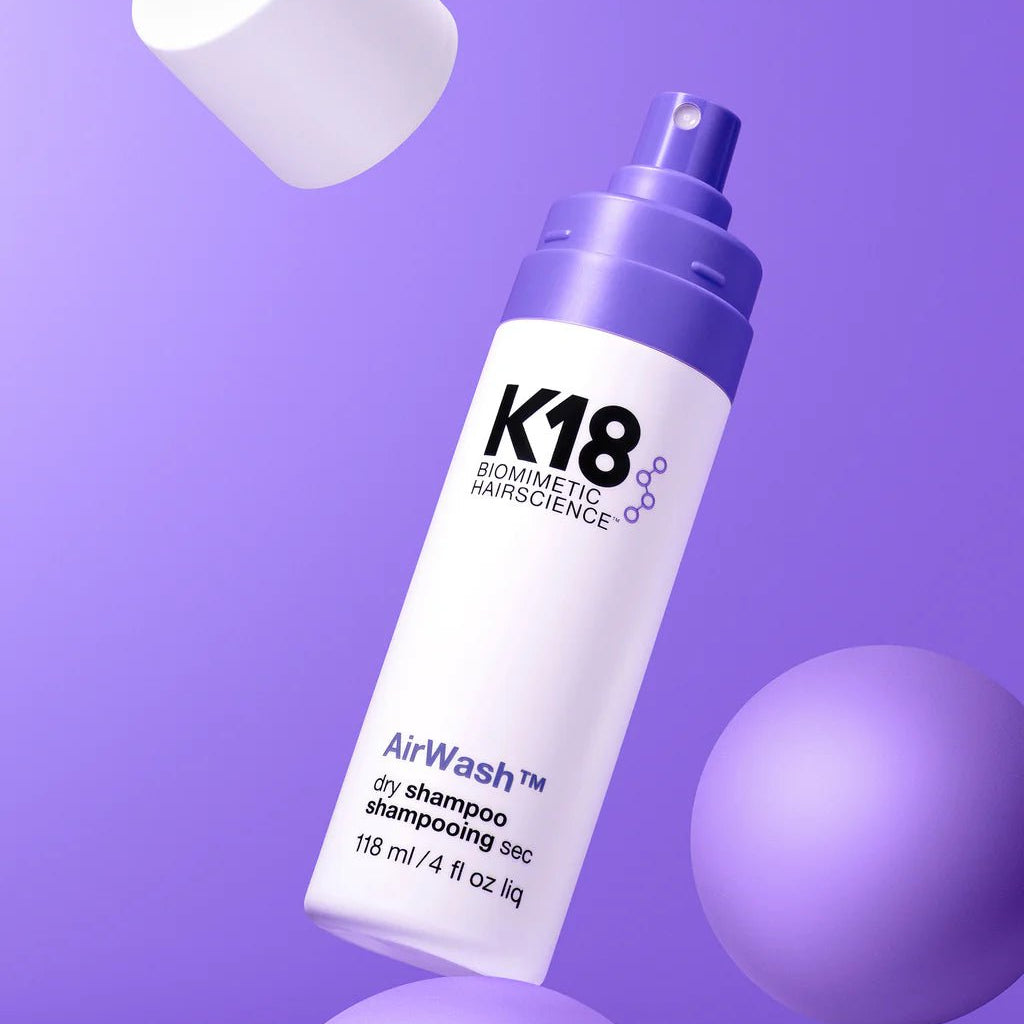 A close-up of a K18 Hair Repair AirWash Dry Shampoo bottle with a purple spray nozzle is set against a purple background, accompanied by two spheres and a detached white cap. The bottle, which reads 118 ml / 4 fl oz liq, features odorBIND™ biotechnology for improved scalp health.