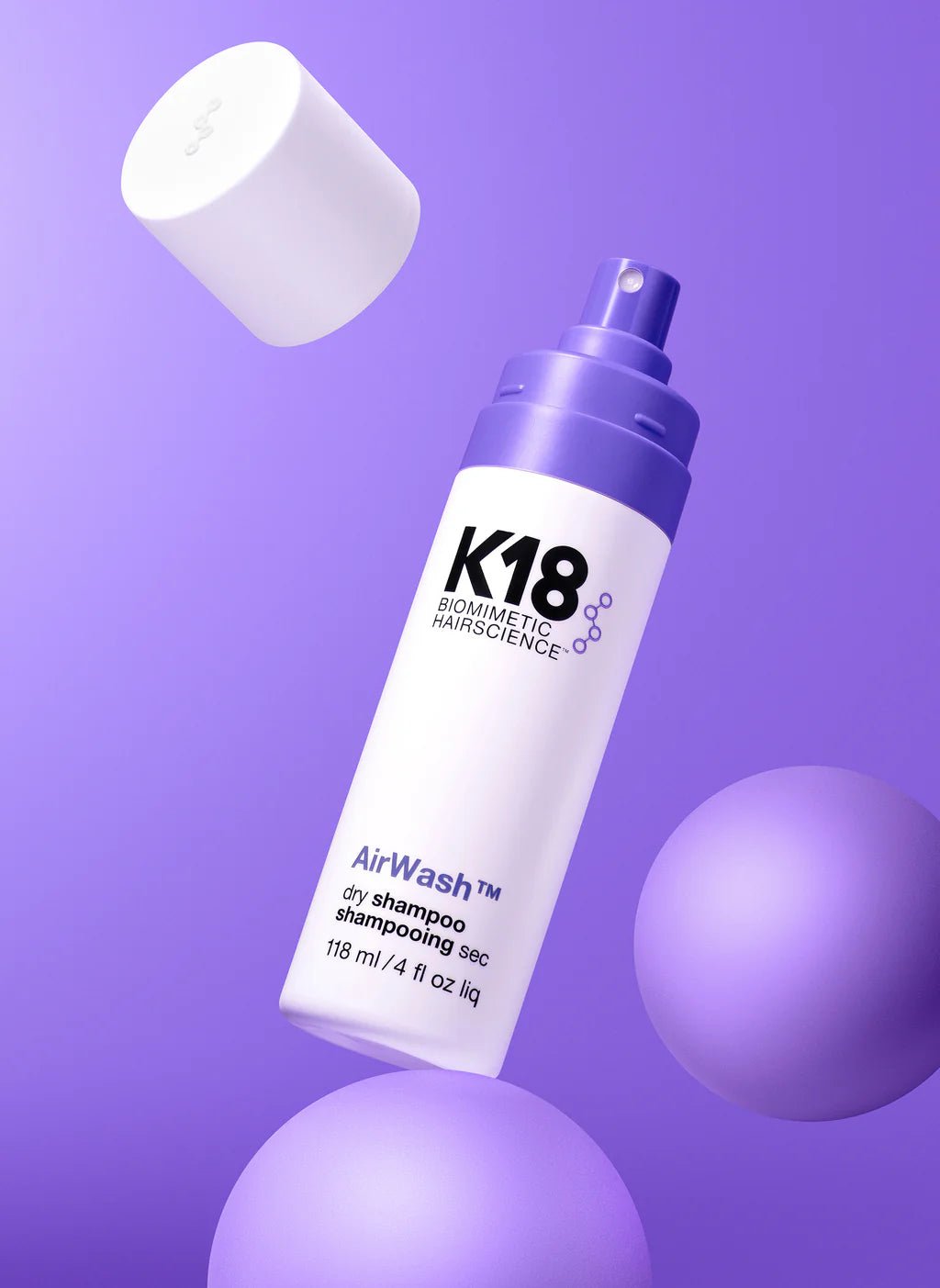 A close-up of a K18 Hair Repair AirWash Dry Shampoo bottle with a purple spray nozzle is set against a purple background, accompanied by two spheres and a detached white cap. The bottle, which reads 118 ml / 4 fl oz liq, features odorBIND™ biotechnology for improved scalp health.