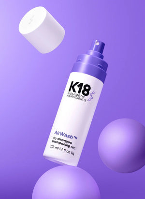 A bottle of K18 Hair Repair K18 AirWash Dry Shampoo with a detached cap is displayed against a purple background featuring spherical shapes.