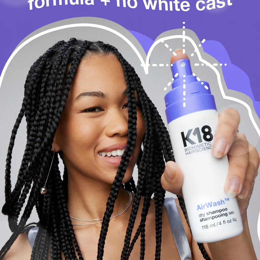 A woman smiling and holding a bottle of K18 AirWash Dry Shampoo, a non-aerosol, scalp-friendly dry shampoo featuring odorBIND™ biotechnology from K18 Hair Repair. The text on the image highlights its formula that leaves no white cast and promotes scalp health.