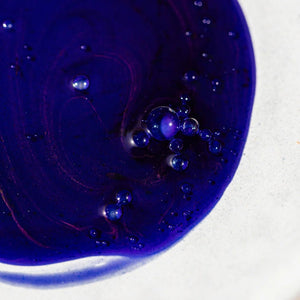A close-up of Oway Bluemoon Hair Bath, an illuminating anti-yellow shampoo for cool blonde, white, and grey hair, shows dark blue liquid with a shiny texture and small bubbles spread on a white background.