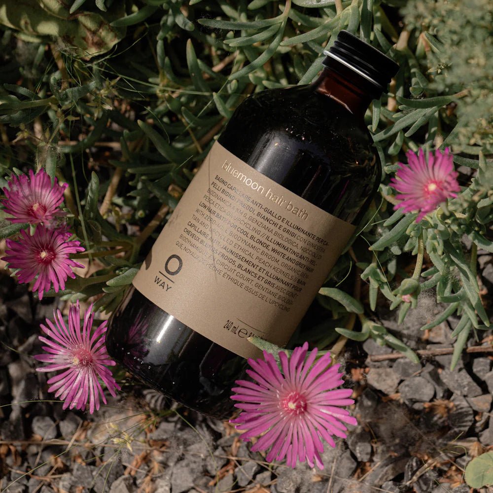 A dark glass bottle labeled "Oway Bluemoon Hair Bath" rests among green plants and pink flowers—an illuminating anti-yellow shampoo by Oway, designed to neutralize brassiness for cool blonde, white, and grey hair.