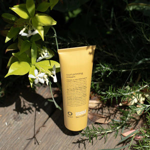 A yellow tube of Oway Curl Priming Cream | Moisture-Rich Curl Primer for Frizz-Free Styling stands upright on a wooden surface amid green foliage and small white flowers, bathed in sunlight. Crafted with Marula Oil, it nourishes and defines curls.