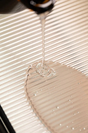 A close-up of Oway Curly Potion | Curl-Defining Serum for Soft, Shiny Curls being poured onto a grooved, light surface, as the clear liquid pools—showcasing Oway’s frizz control and curl-enhancing benefits.