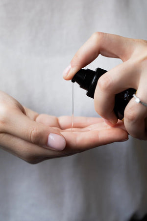A person dispenses Oway Curly Potion | Curl-Defining Serum for Soft, Shiny Curls from a black pump bottle onto their palm. Wearing a light top, they prepare for smooth styling and frizz control; the background is softly blurred.