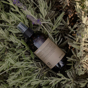 A brown bottle labeled "Oway Flowerfall | Aromatherapeutic Scalp and Mind Balancing Mist" sits on lush green lavender foliage. The product is from Oway, designed to balance scalp and mind with its aromatherapeutic formula.
