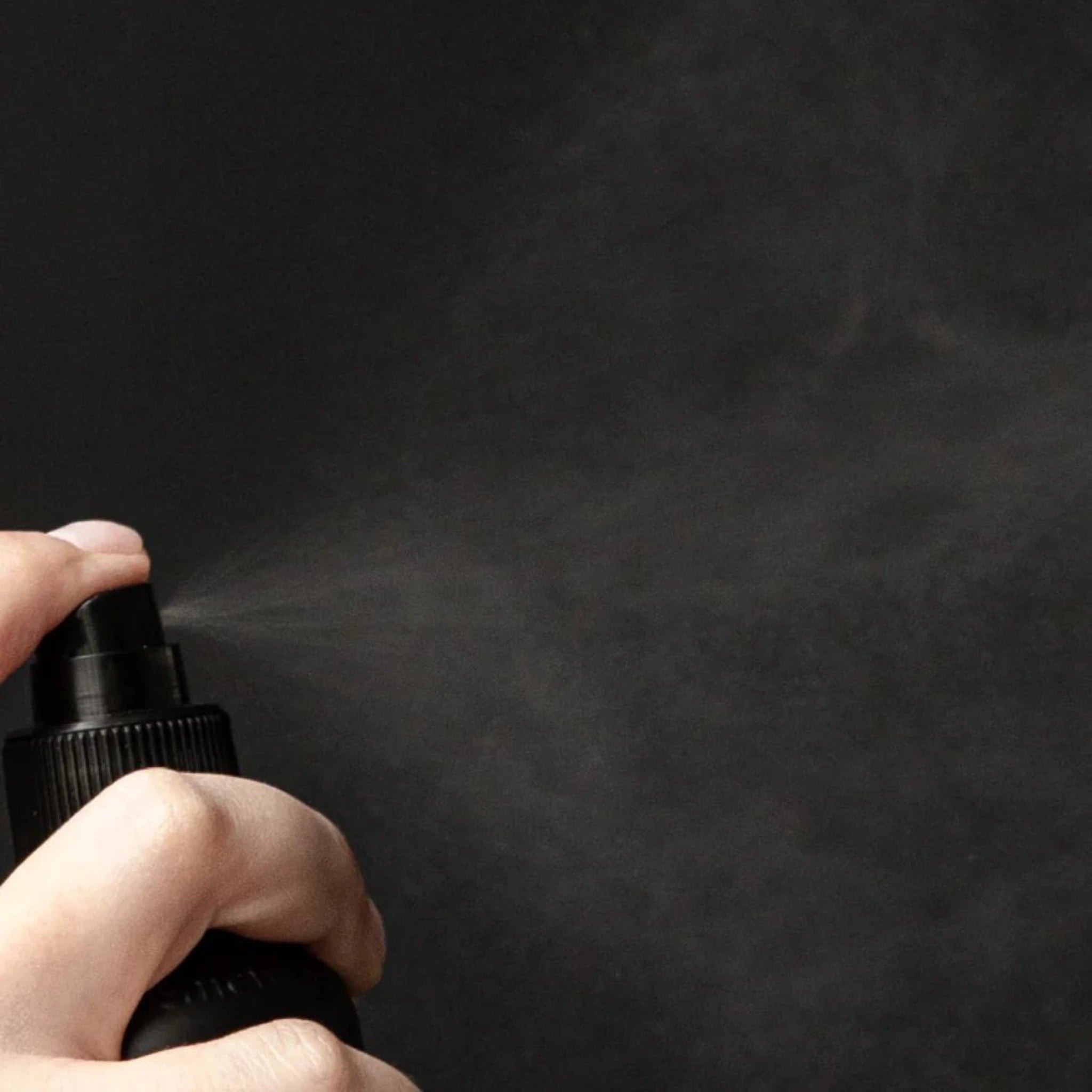 A hand presses the nozzle of a black Oway Flowerfall | Aromatherapeutic Scalp and Mind Balancing Mist bottle, releasing a fine spray against a dark background.