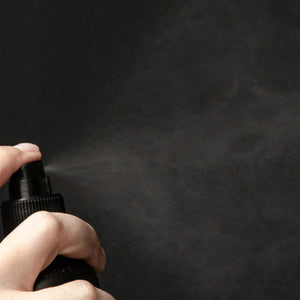 A hand presses the nozzle of a black Oway Flowerfall | Aromatherapeutic Scalp and Mind Balancing Mist bottle, releasing a fine spray against a dark background.