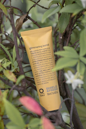 A yellow Oway Glimmering Bond tube—Bonding Shine Treatment for Damaged, Dull Hair—stands upright among green leaves and white flowers. The label displays product details in both Italian and English.