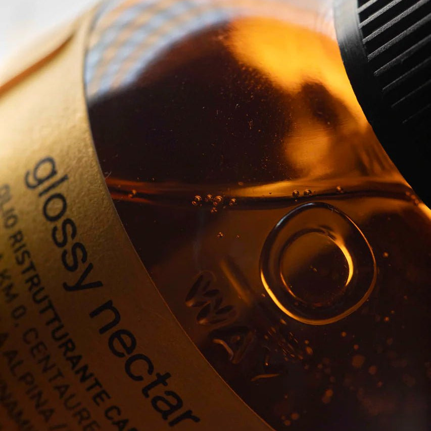 A close-up of a brown Oway Glossy Nectar | Rebuilding Oil Serum for Shine and Hair Strength bottle, showing the cap, product name, embossed measurement markings, and frizz control benefits on the label.