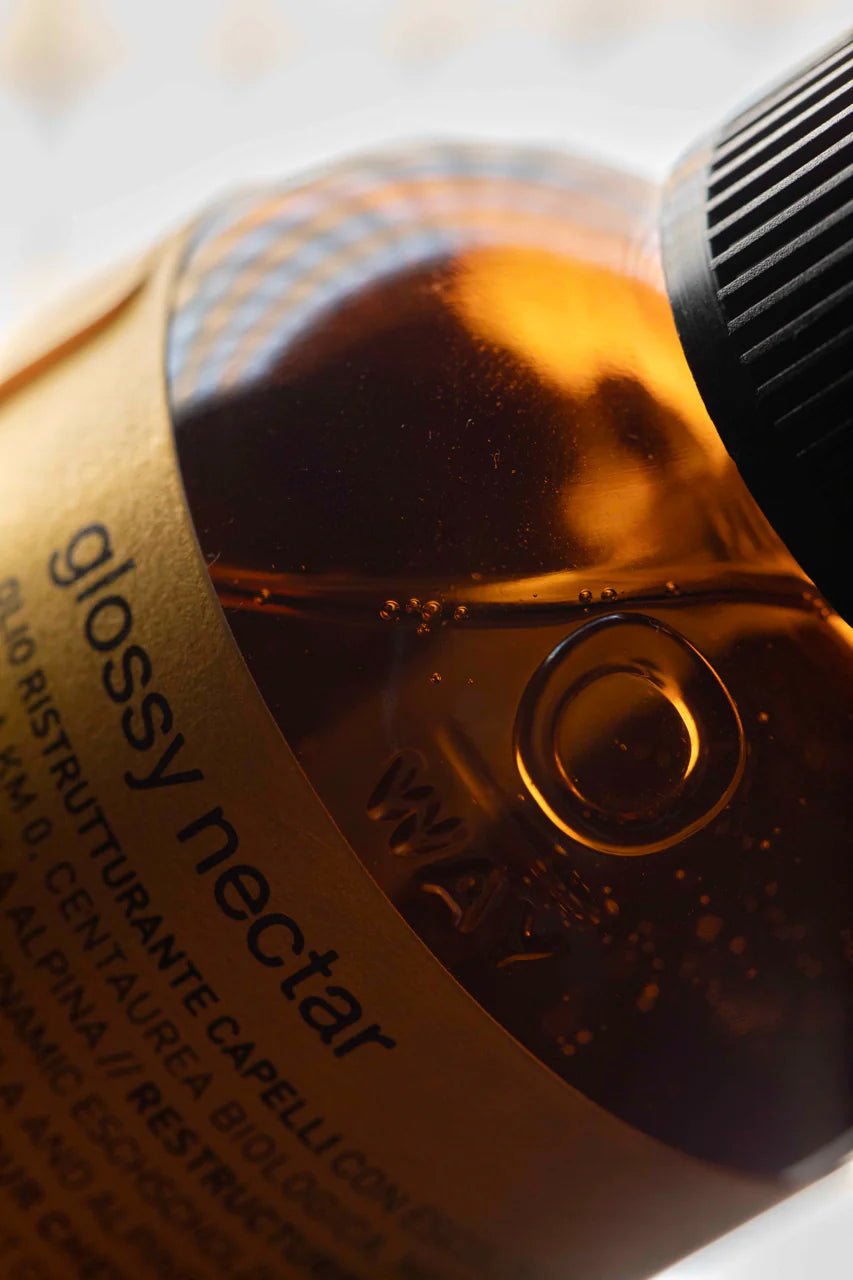 A close-up of a brown Oway Glossy Nectar | Rebuilding Oil Serum for Shine and Hair Strength bottle, showing the cap, product name, embossed measurement markings, and frizz control benefits on the label.