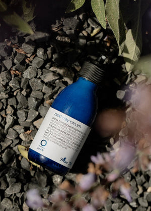 A bottle of Oway Next Day Cream | Soft Styling Cream for Next-Day Texture Revival with a black pump sits on dark gravel among leafy plants and blurred purple flowers, ready to refresh your hair’s texture.