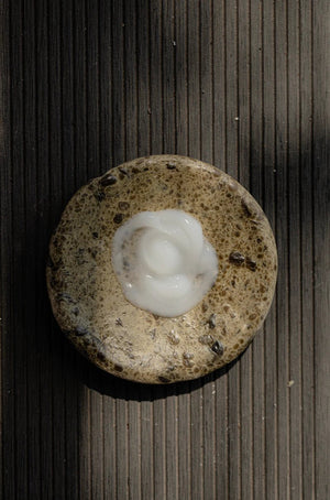 A touch of Oway Next Day Cream | Soft Styling Cream for Next-Day Texture Revival is elegantly swirled atop a textured, round brown stone on a striped surface, capturing the essence of Oway’s next-day texture revival.