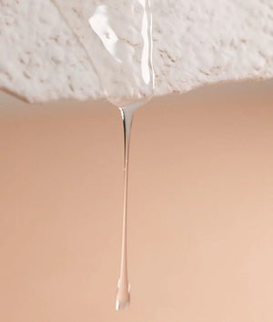 A close-up of a clear liquid drip hanging from a textured white surface above a beige background, evoking the purity and renewal found in Oway Rebuilding Hair Bath, the strengthening shampoo for brittle, weak hair.