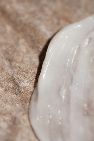 A close-up view of Oway Rebuilding Hair Mask, a white creamy restorative mask for damage and instant repair, spread on a textured brown surface—highlighting its rich texture, ideal for treating chemically treated hair.